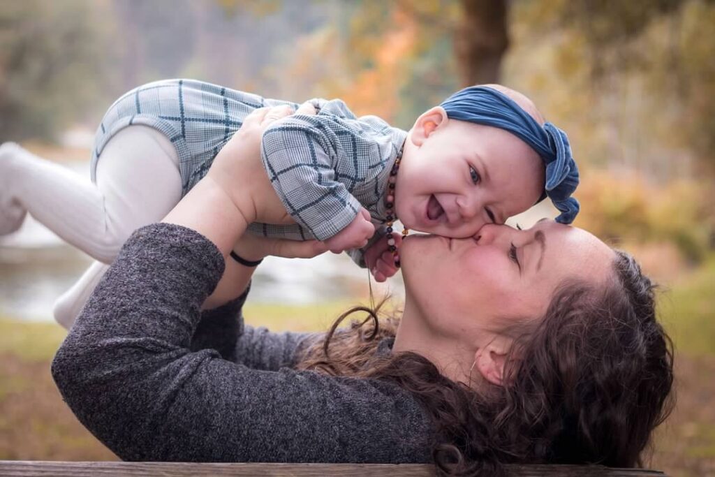 a woman kissing her laughing baby girl on a bench in the forest on a fall day
